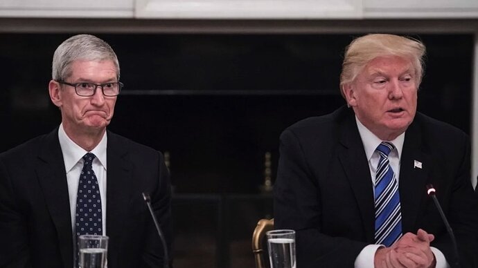 Two men in suits sit at a table, one with glasses and a neutral expression, the other speaking with a serious expression.
